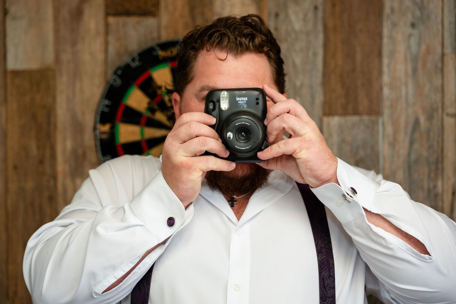 Bearded man in white shirt and suspenders taking a photo with a Fujifilm Instax camera near a dartboard.