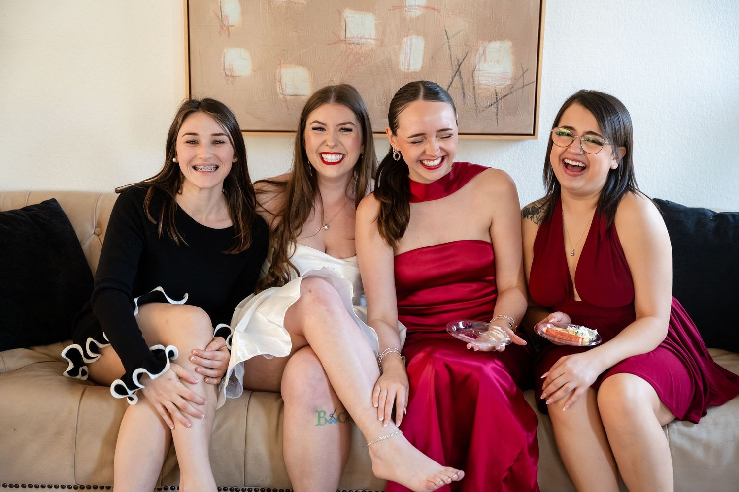 Four smiling women in elegant dresses sitting together on a couch at a formal celebration.