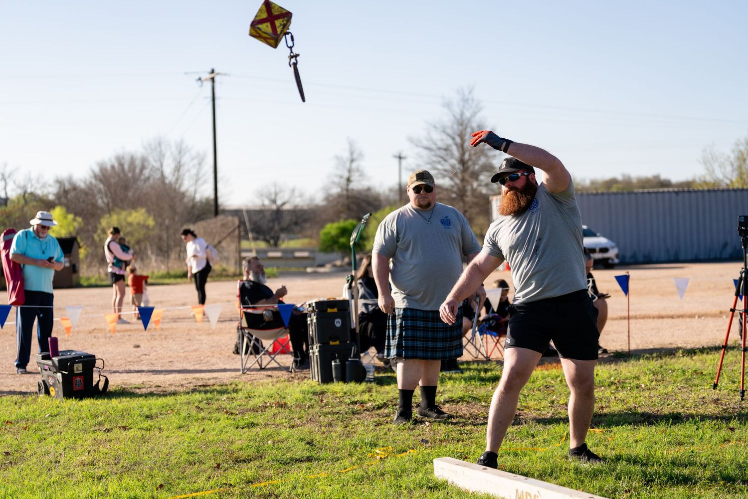 People playing outdoor lawn games on a sunny day, with a man throwing a ball and a kite flying in the background.