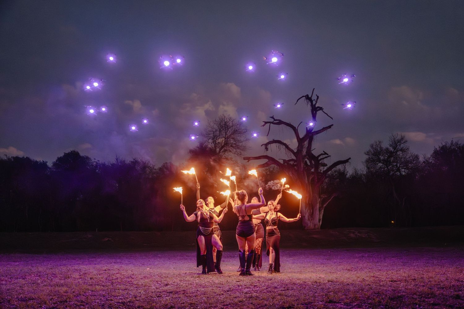 Fire dancers perform at night with sparkling lights above a dramatic dead tree in a purple-lit outdoor setting.