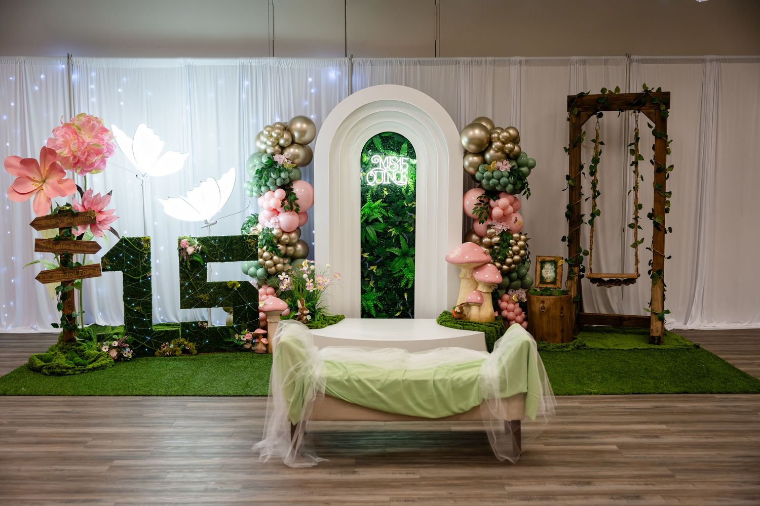 Elegant quinceañera setup with number 15, floral arch, pink blooms, gold balloons, and draped table on artificial grass.