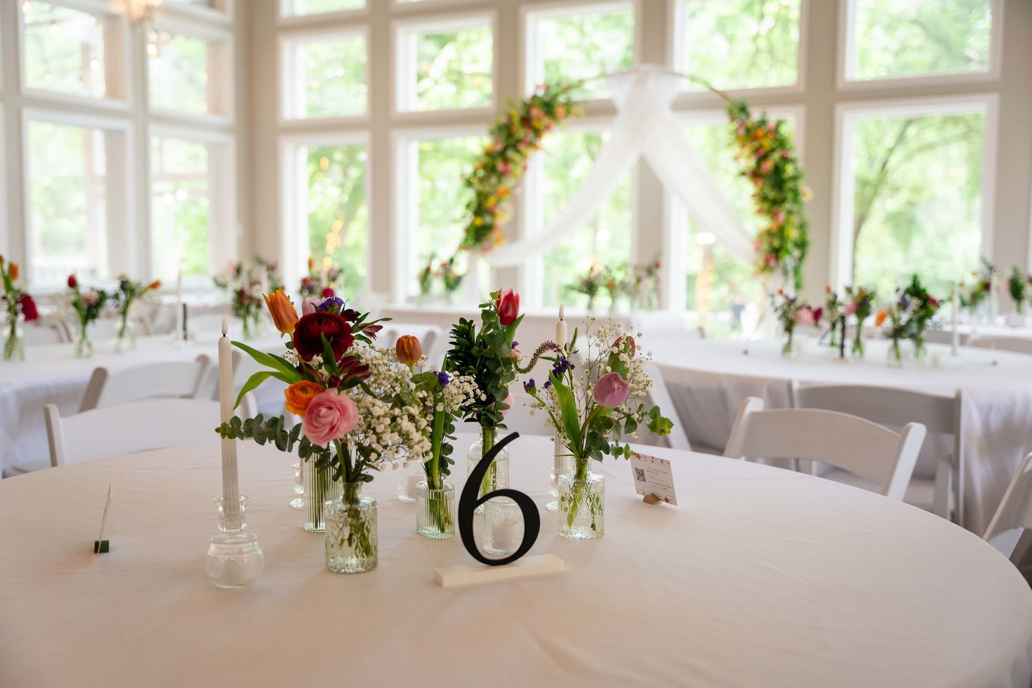 Elegant wedding reception hall with floral centerpieces, table number six, white linens, and a floral arch backdrop by bright windows.