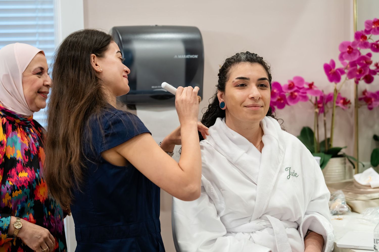 Woman in white robe receiving makeup application in spa, with two women watching near pink orchids.