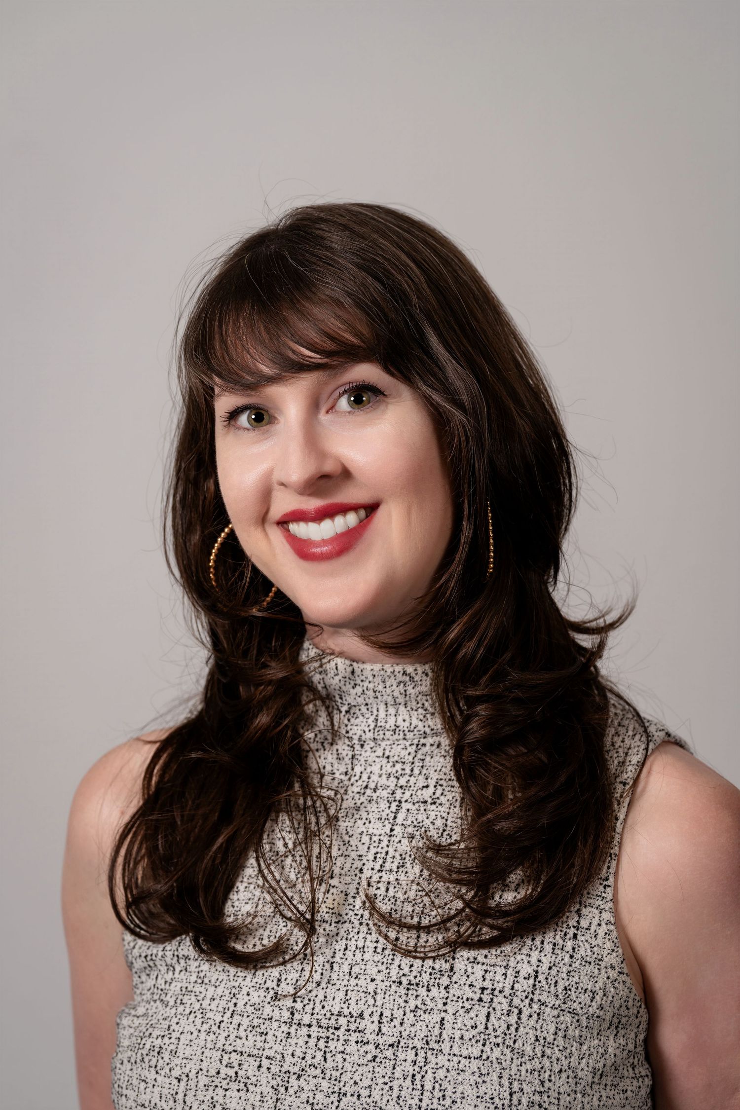 Smiling brunette woman with hoop earrings wearing a gray tweed sleeveless top against a light gray background.