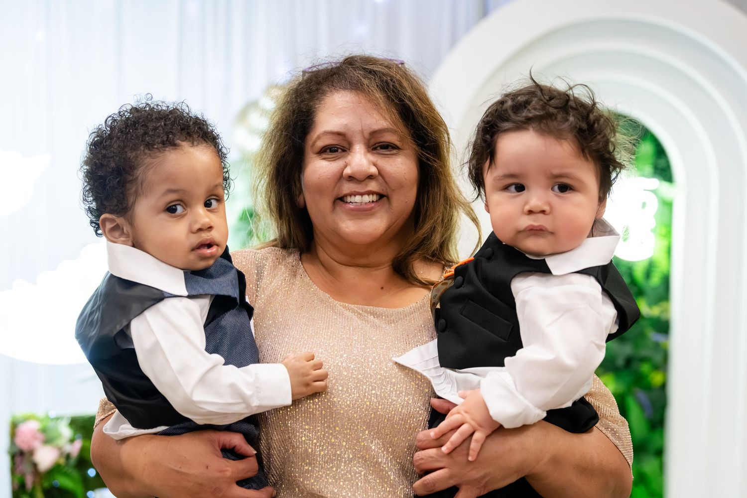 A smiling woman holds two dressed-up baby boys in formal vests at an elegant event.