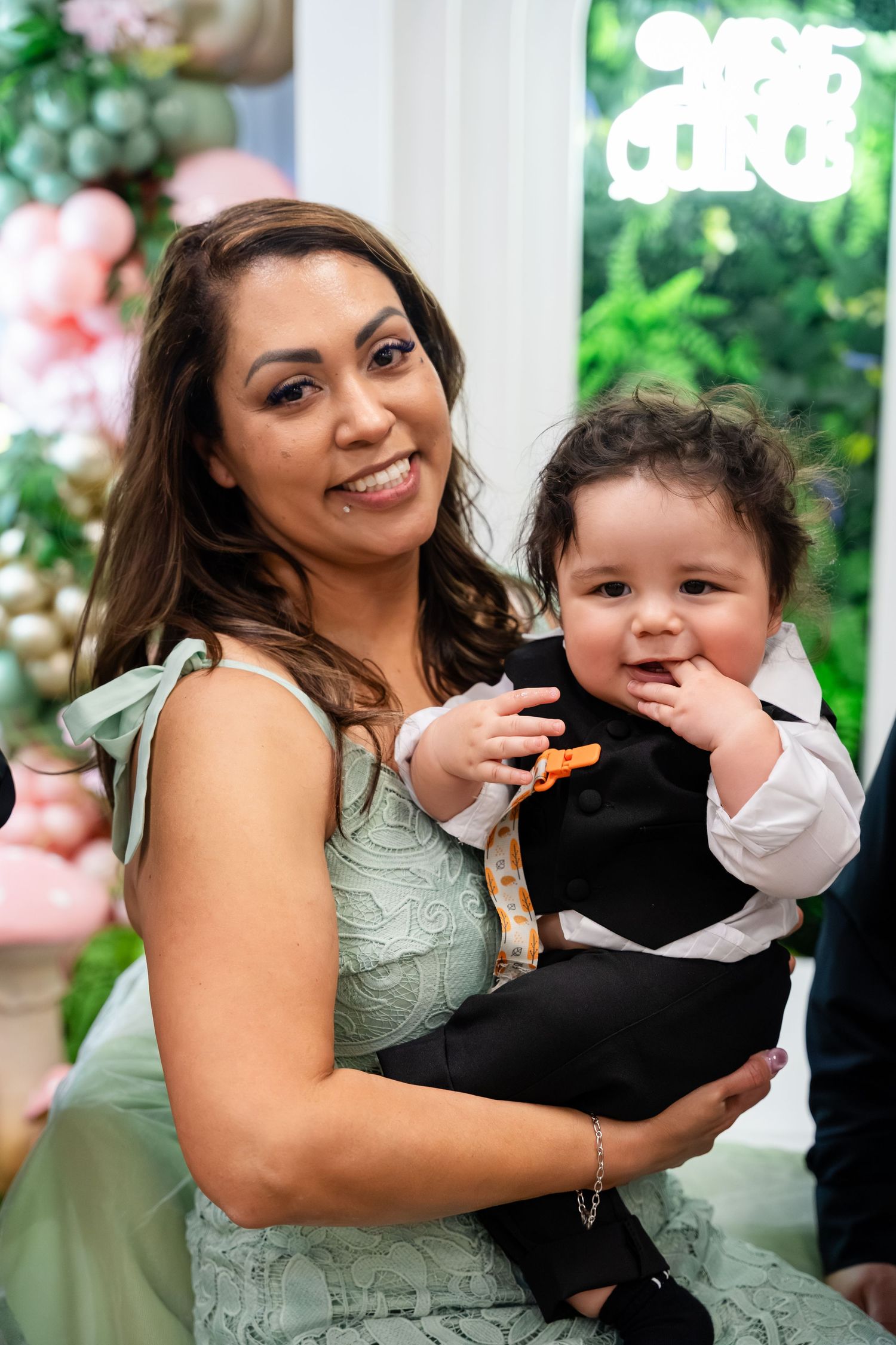 A smiling woman in a sage green dress holds a baby boy in a tuxedo at a festive celebration with balloon decorations.
