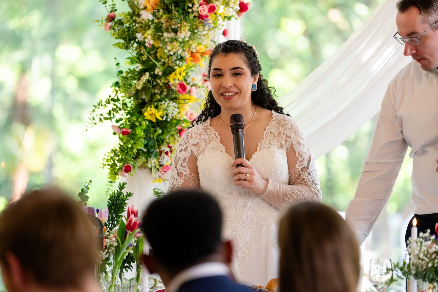 Bride in lace wedding dress speaks into microphone at wedding reception with floral backdrop.