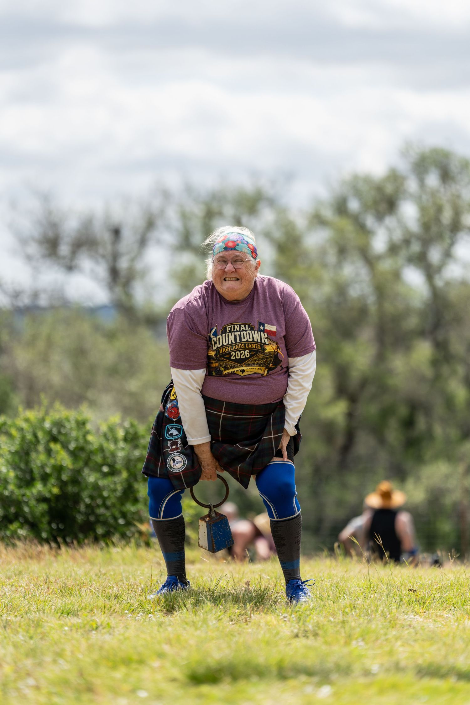 Competitor in a purple shirt and kilt participates in a Highland Games event on a grassy field.