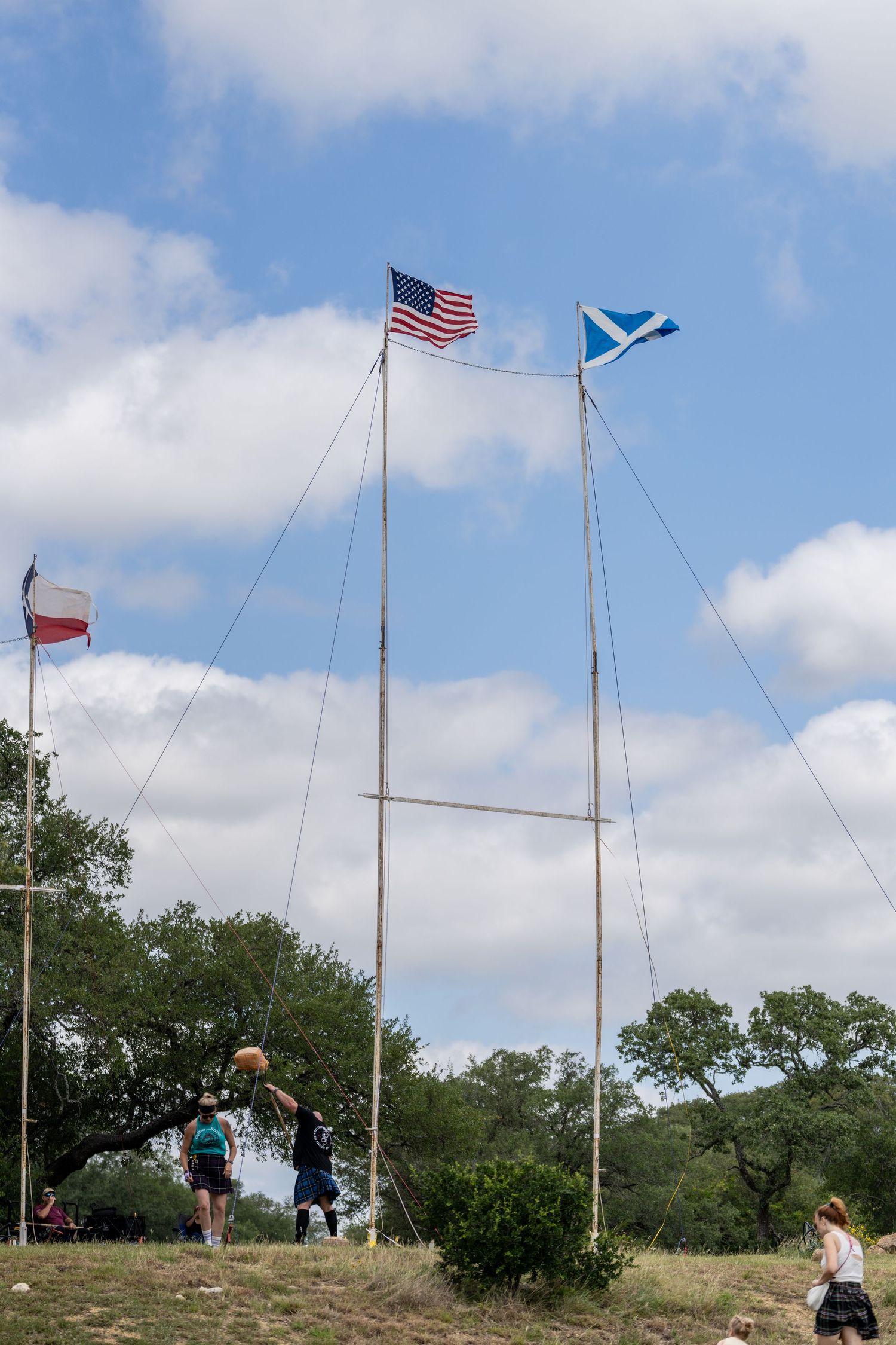 People gather at outdoor Highland Games event with American, Scottish, and Texas flags flying on tall poles against blue sky.