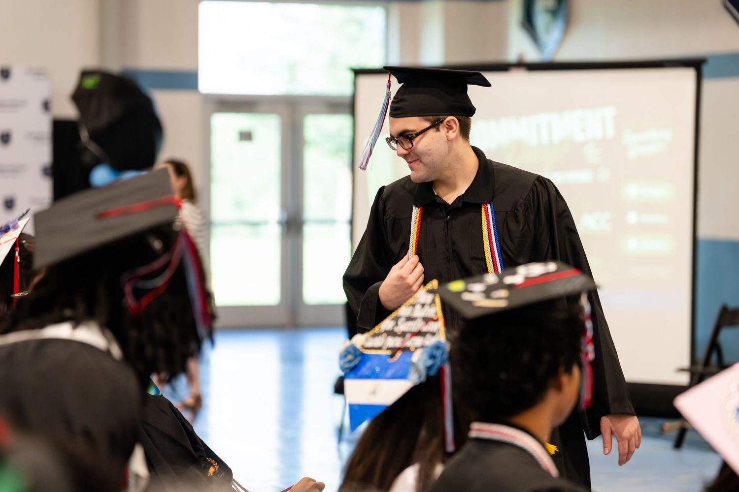 Graduate in cap and gown holding a diploma at a high school graduation ceremony indoors.