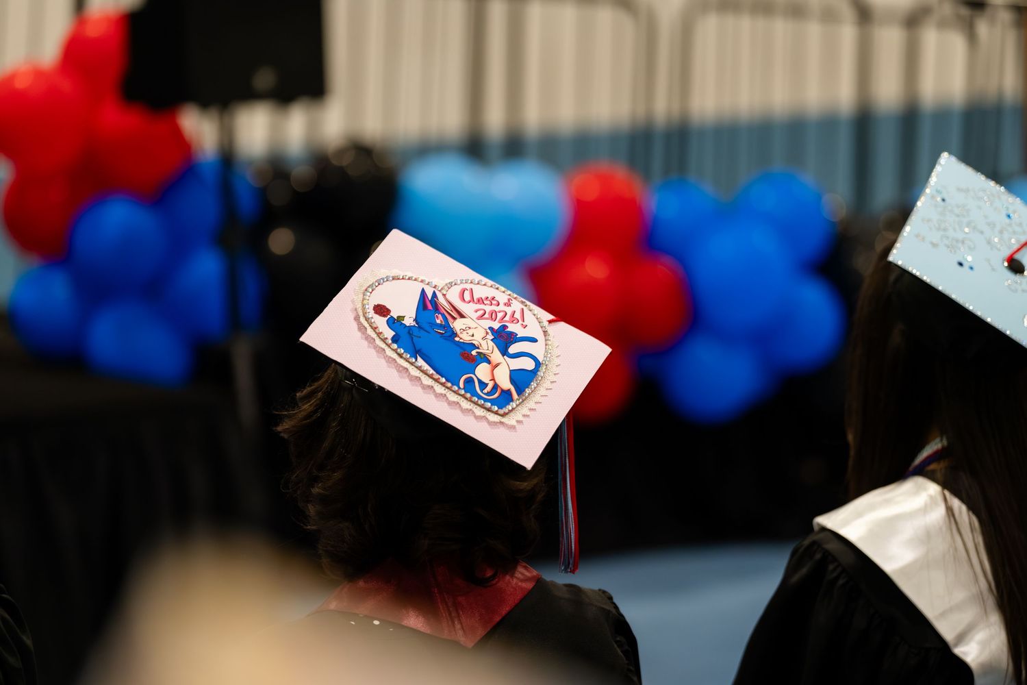 Graduation cap decorated with Disney design at ceremony with red and blue balloons in background.