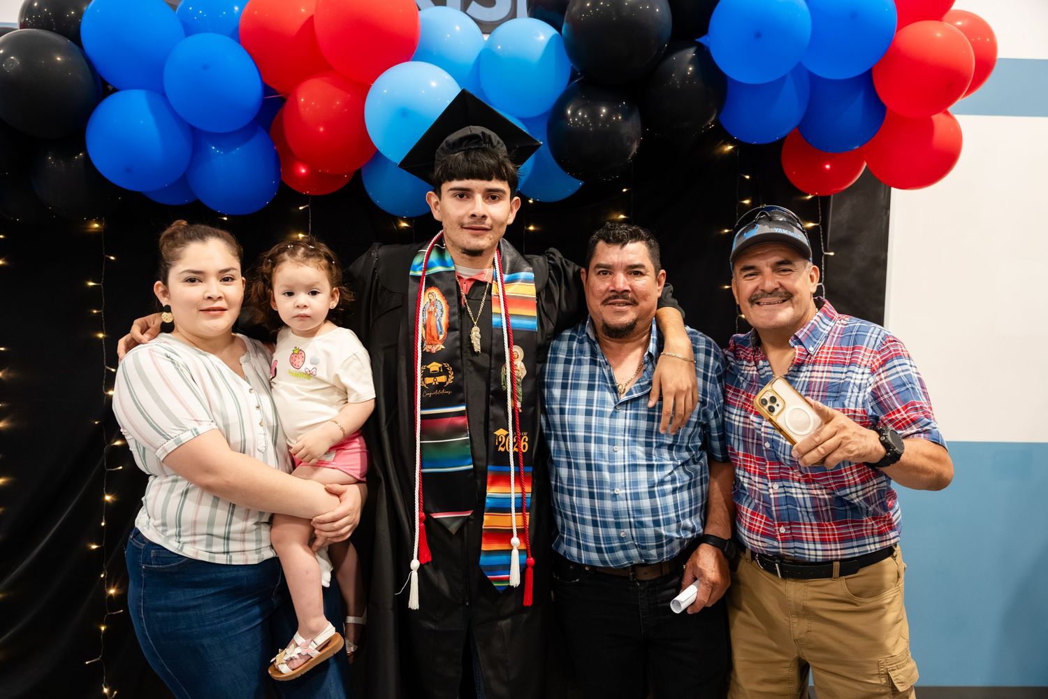 Family celebrating graduation with graduate in cap and gown, posing under red, blue, and black balloon arch.