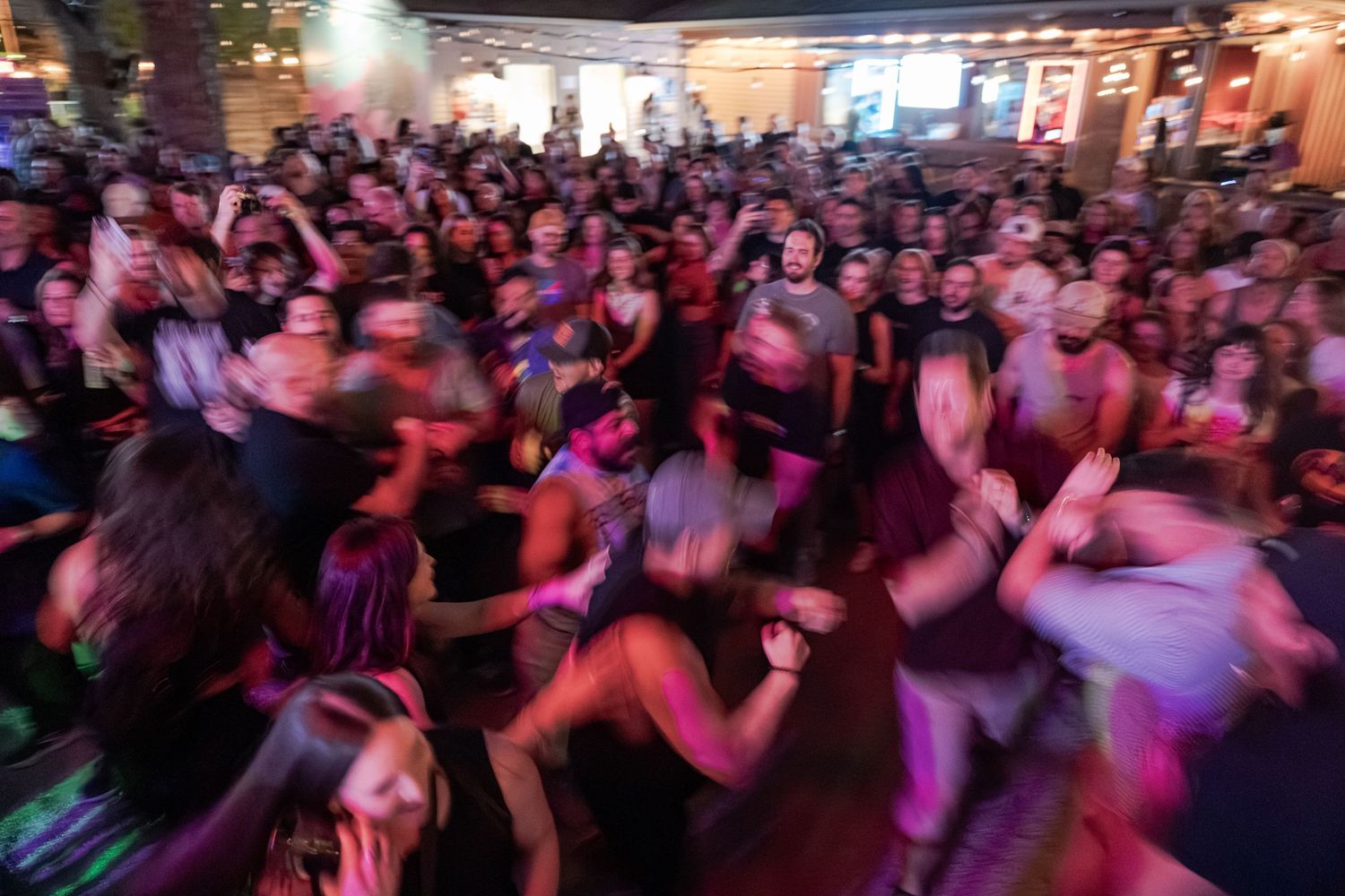 A large energetic crowd of people dancing at an outdoor concert or festival event under colorful lights.
