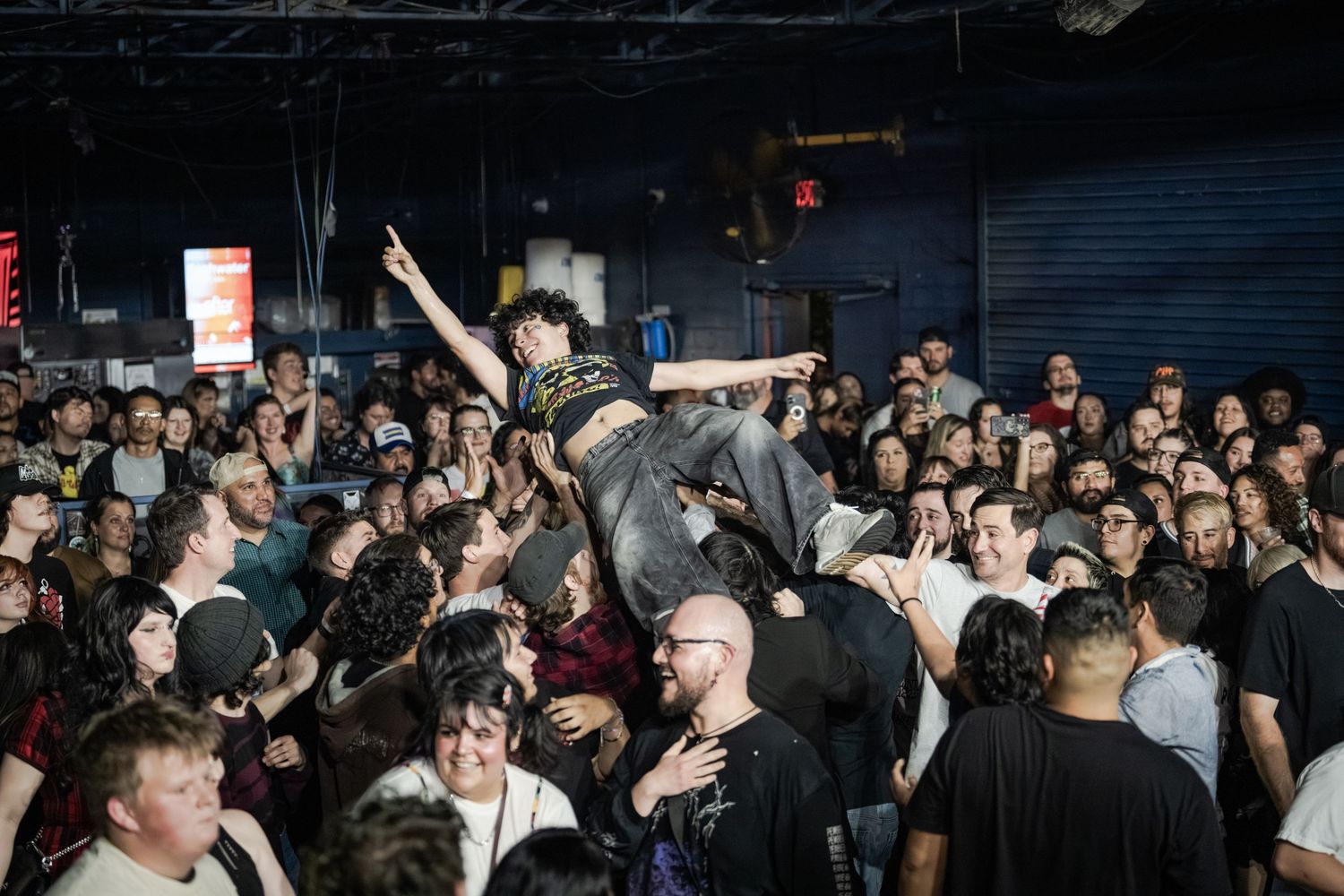 Crowd surfer raised above dense concert crowd in dark venue, arms outstretched, at energetic punk rock show.