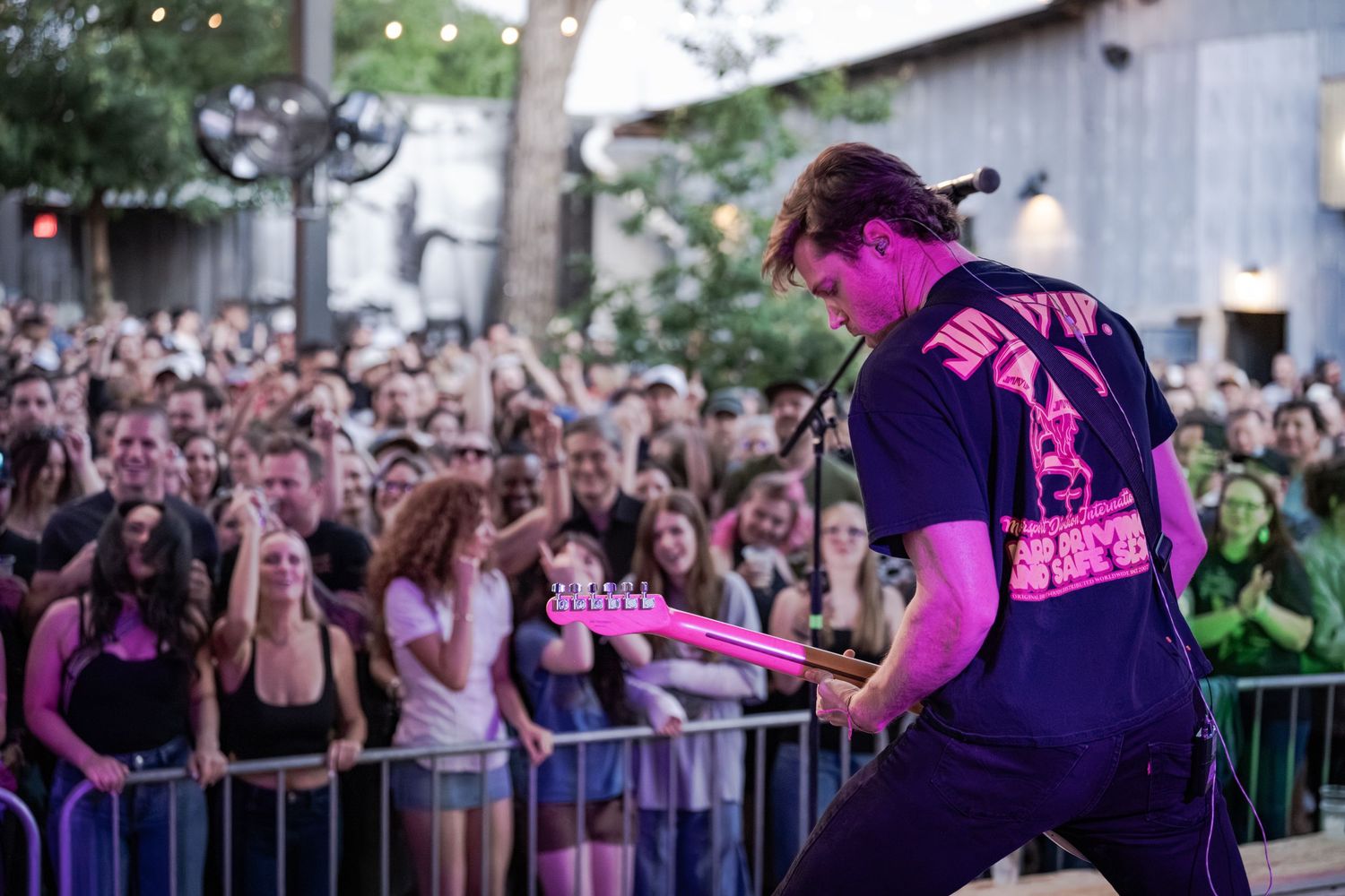 Guitarist in black graphic tee performs on outdoor stage before large crowd at energetic live music concert.