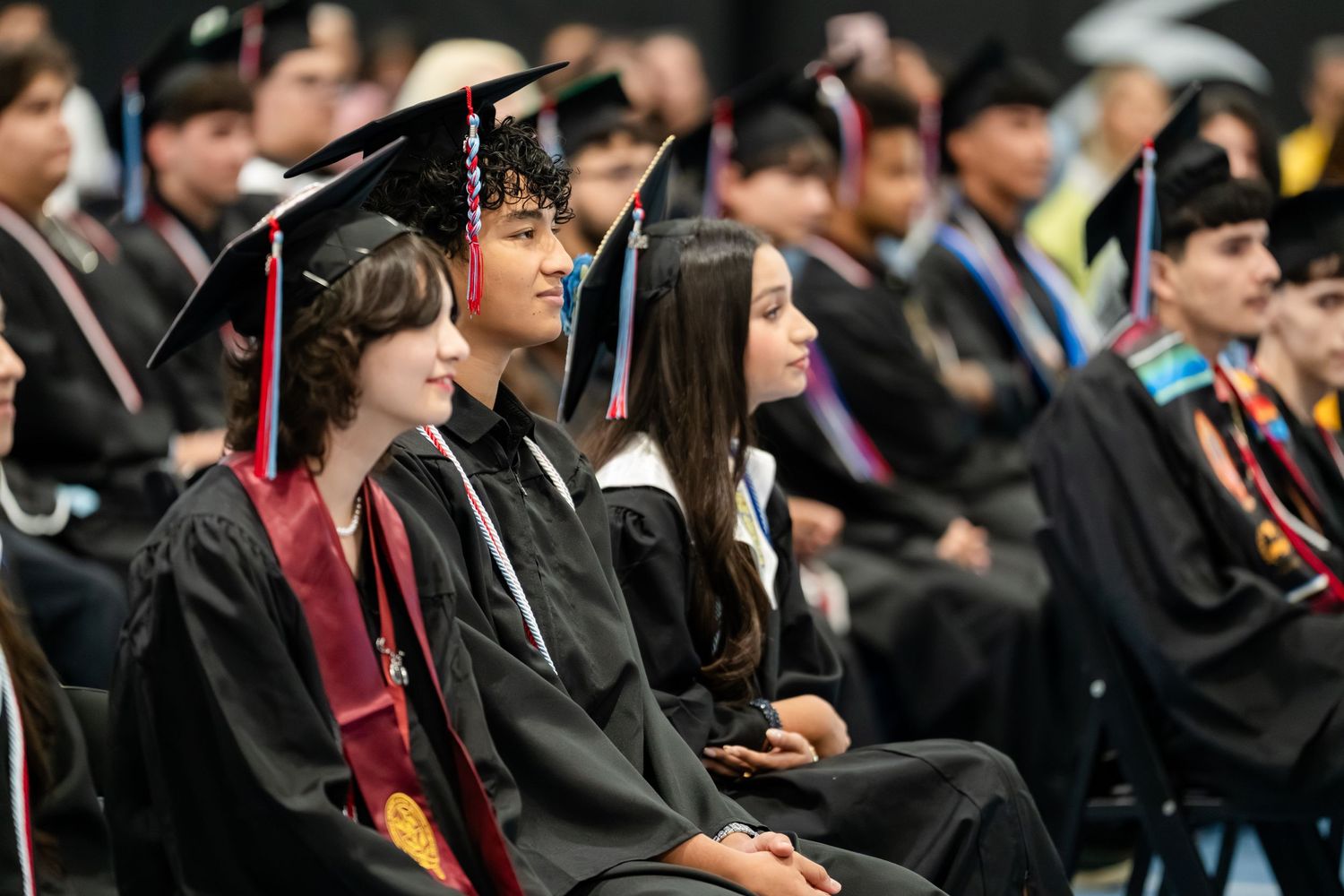 Students in graduation caps and gowns seated at a commencement ceremony, smiling and celebrating their achievement.