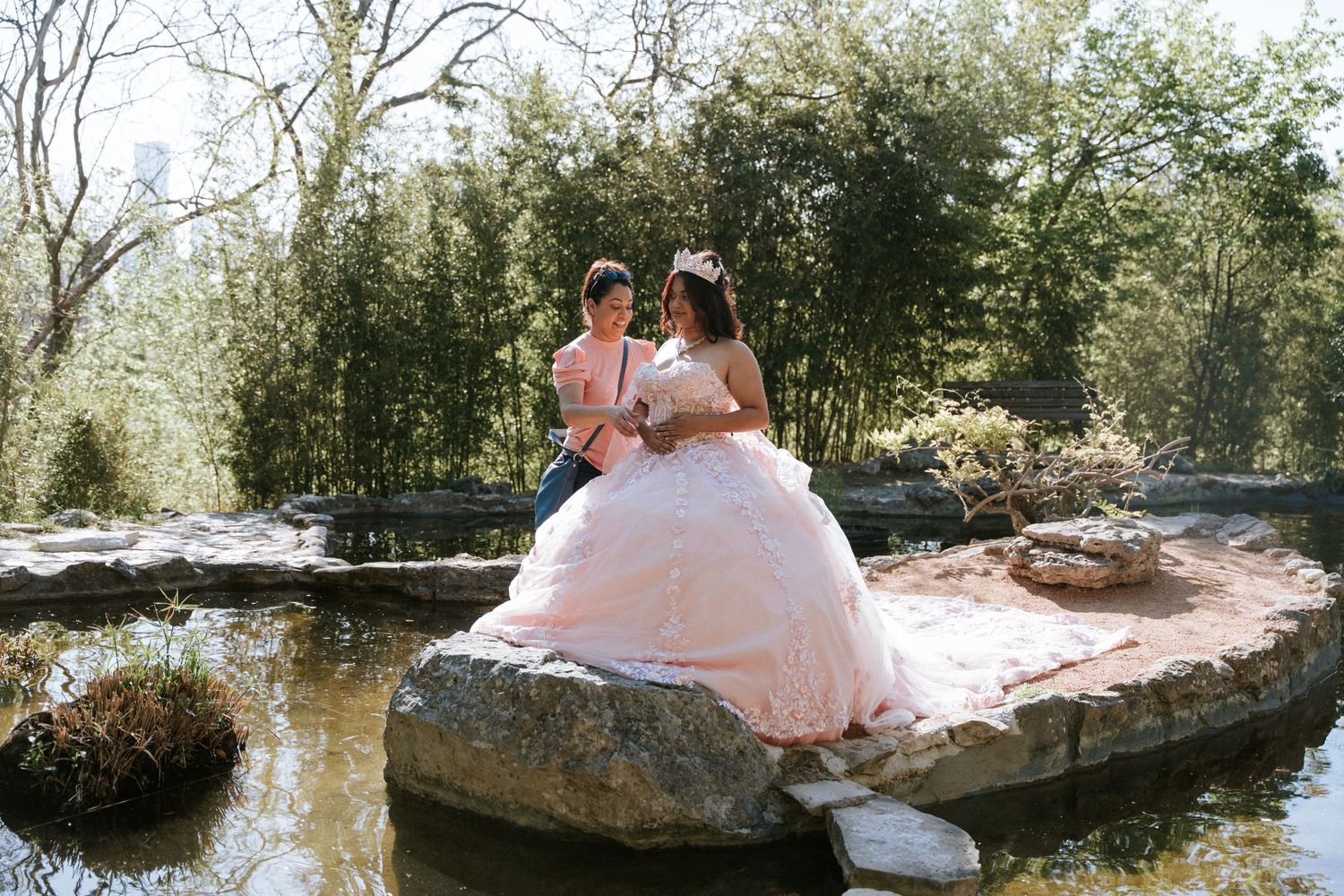 A bride in a pink ball gown and groom sit romantically on rocks by a pond in a lush garden setting.