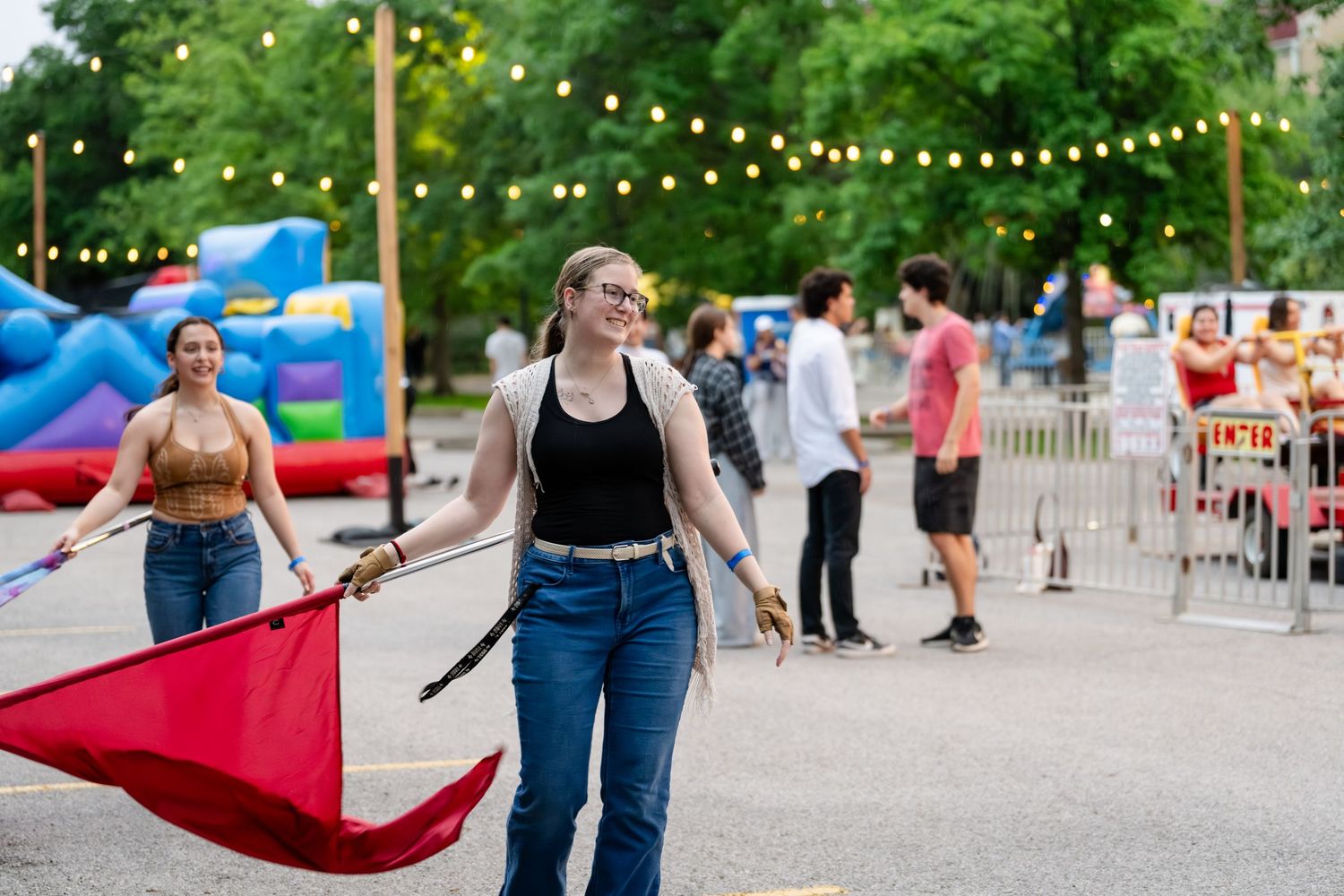 Young woman smiling and holding a red flag at an outdoor carnival event with string lights and inflatable attractions.