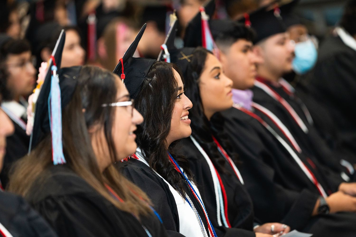 Graduates wearing black caps and gowns with red and white stoles sit in rows during a commencement ceremony.