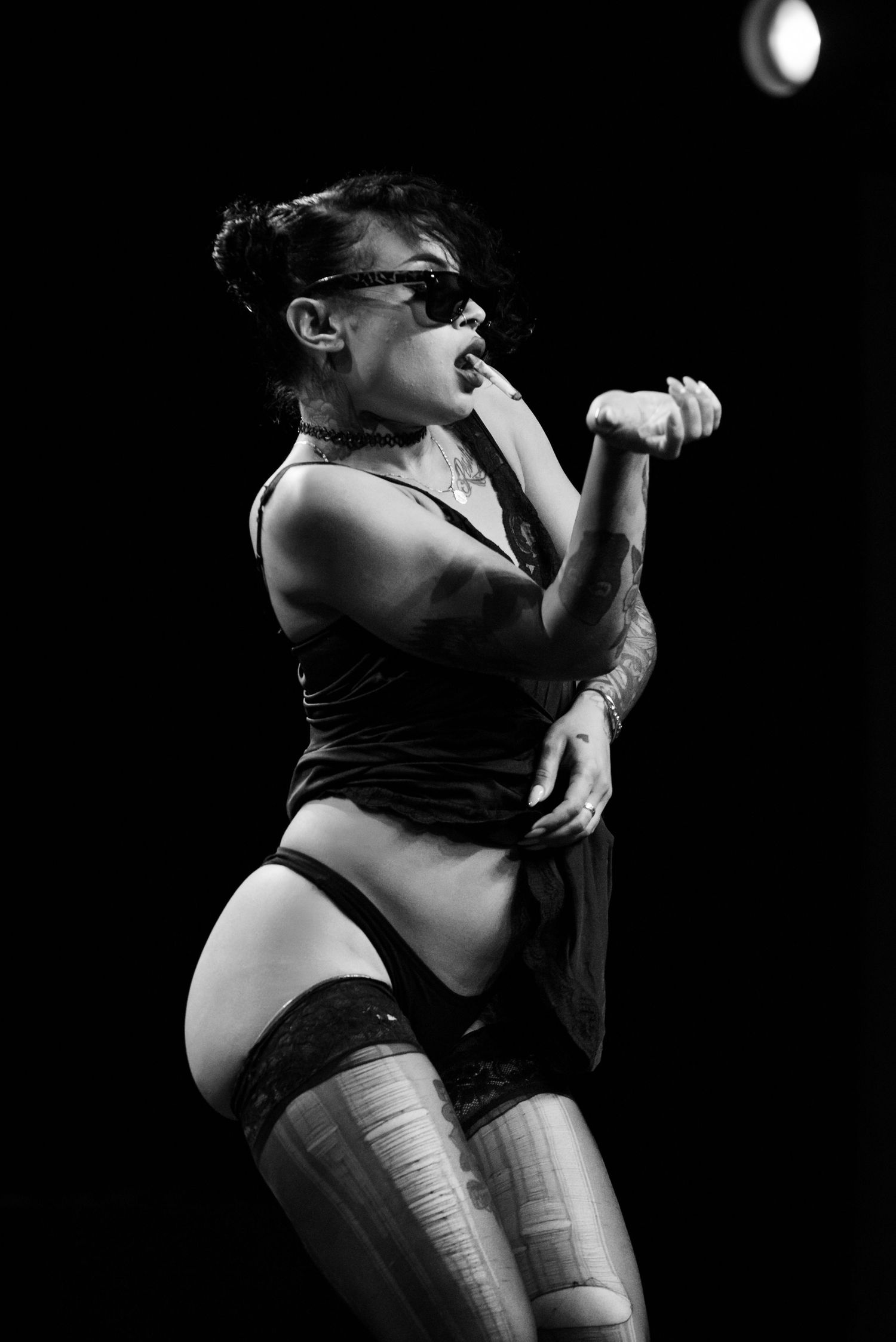 Black and white photo of a performer in sunglasses and lingerie singing on stage with dramatic lighting.