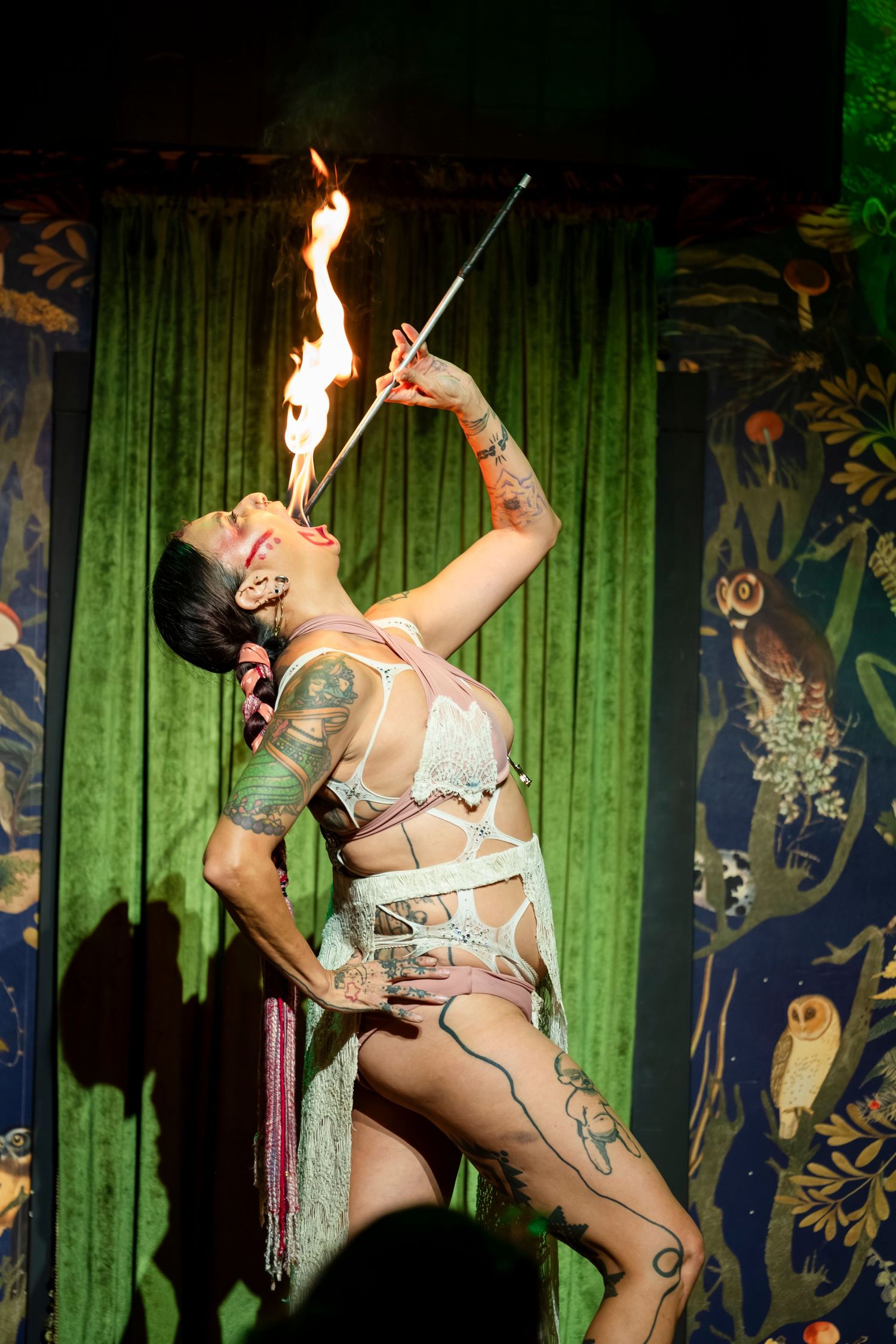 Burlesque performer in silver costume performs sword swallowing act on stage with green curtain backdrop.