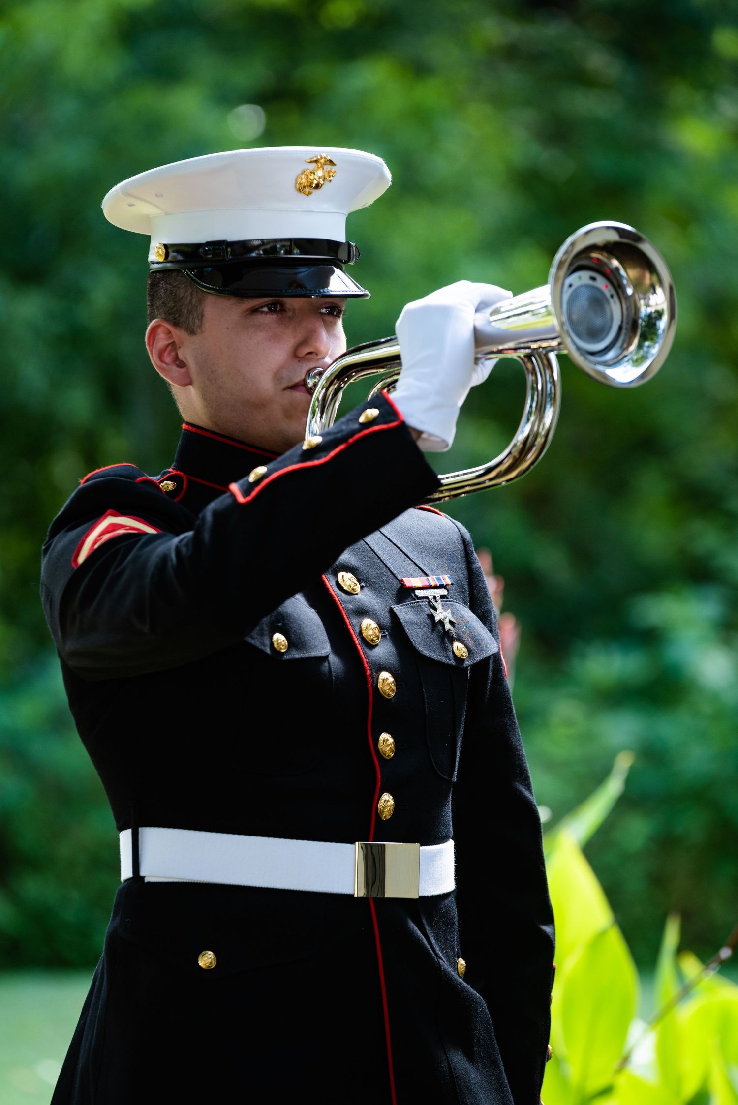 A military bugler in dress uniform plays taps on a ceremonial bugle outdoors with green foliage in the background.