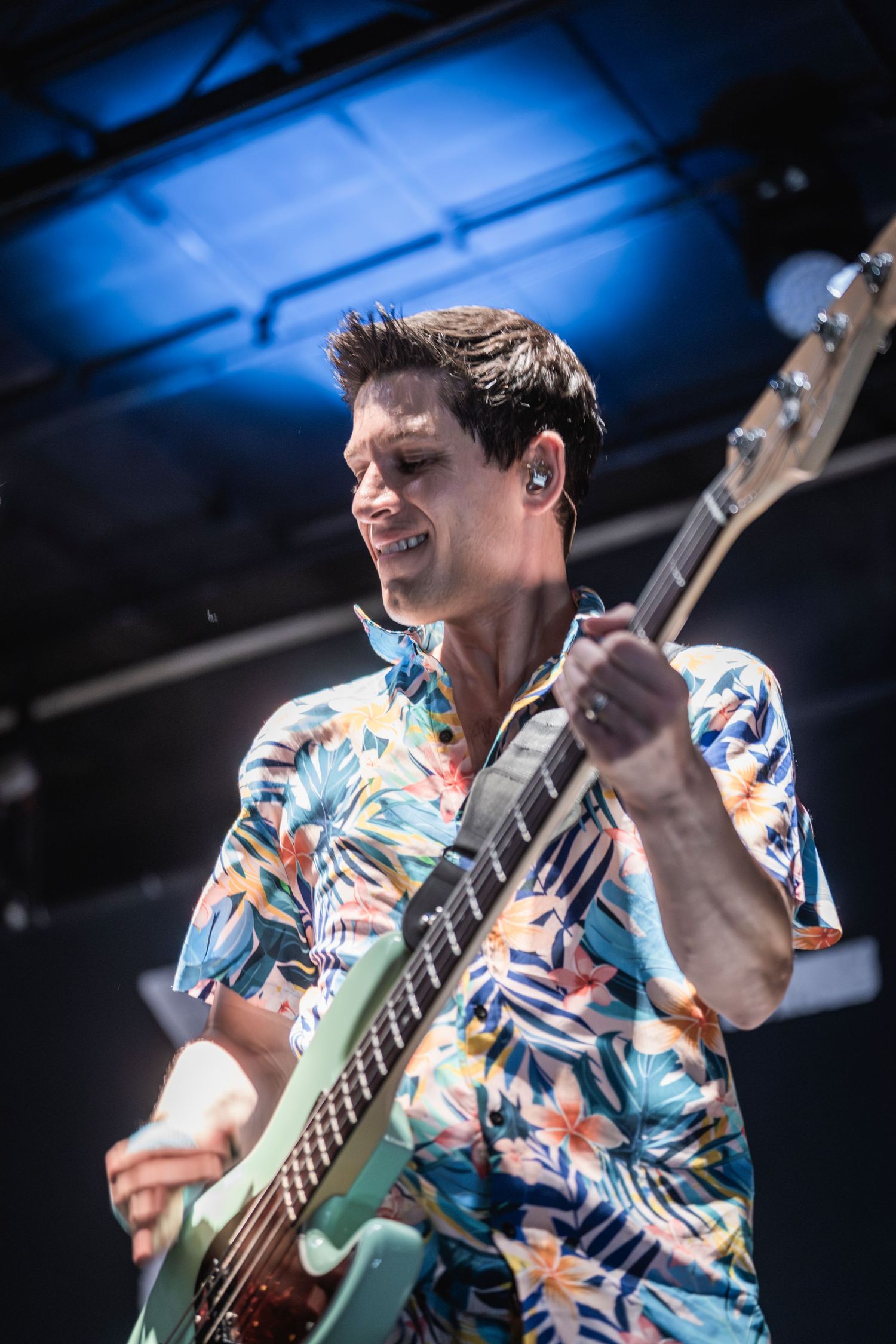 Musician in floral Hawaiian shirt performs energetically on bass guitar against dramatic blue stage lighting.