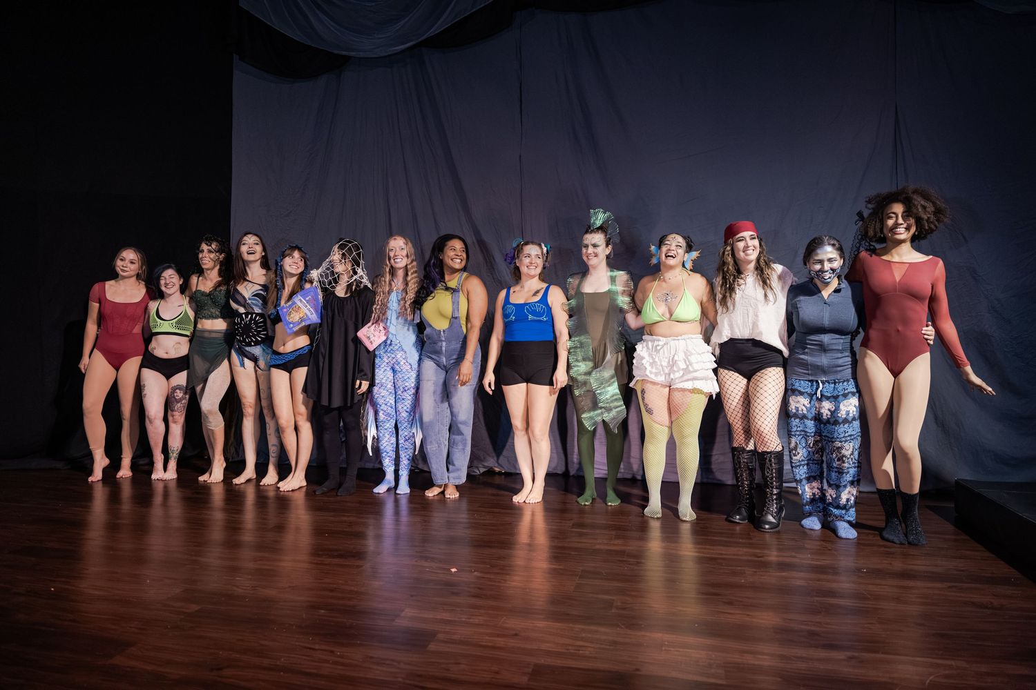 Group of diverse women in colorful lingerie and costumes posing together on a stage with dark backdrop.