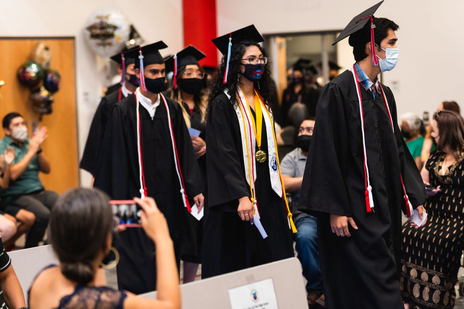 Masked graduates in black gowns and caps walking in procession during ceremony.