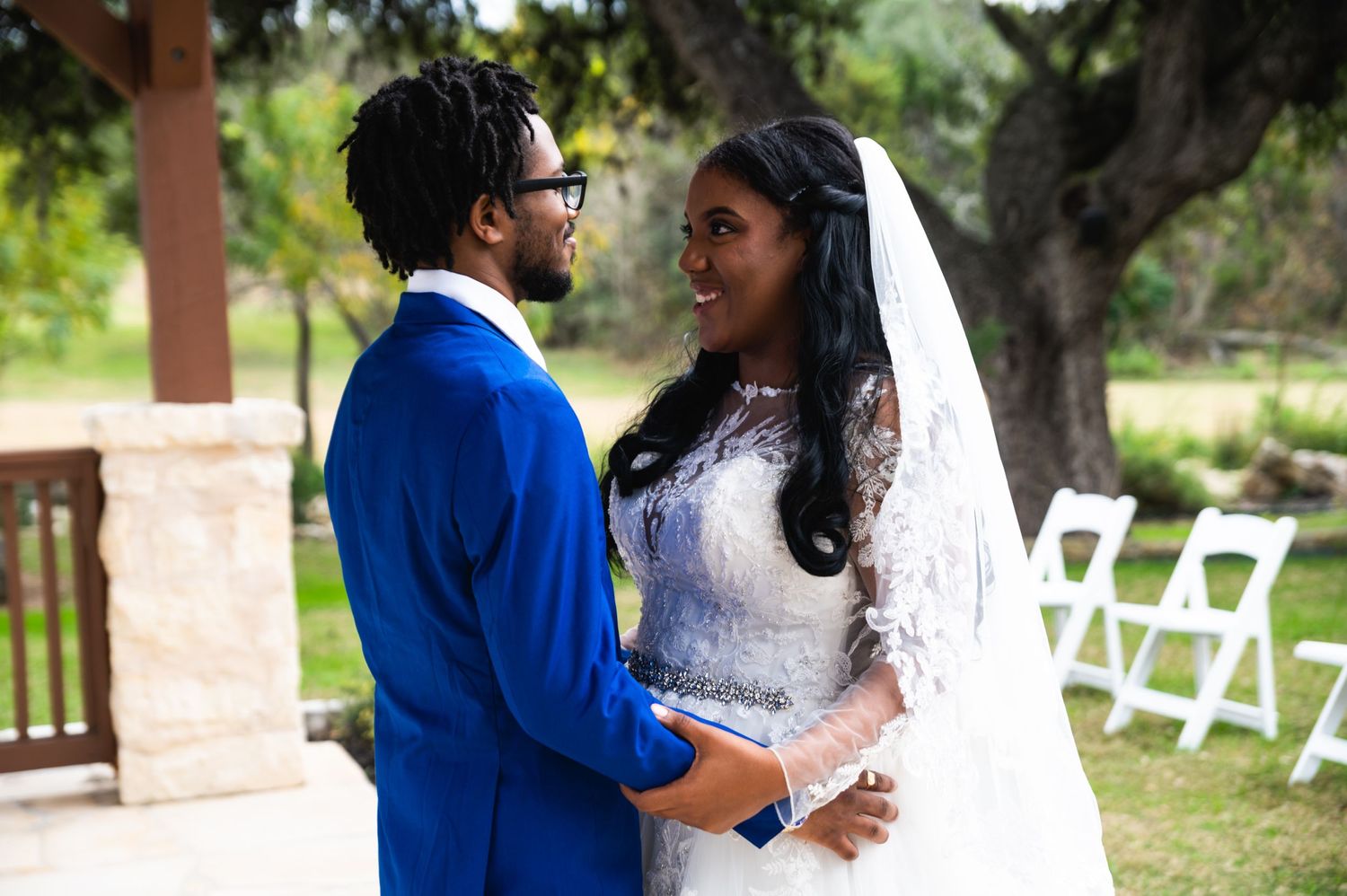 A tender wedding moment captured under a wooden pavilion with white chairs in the background.