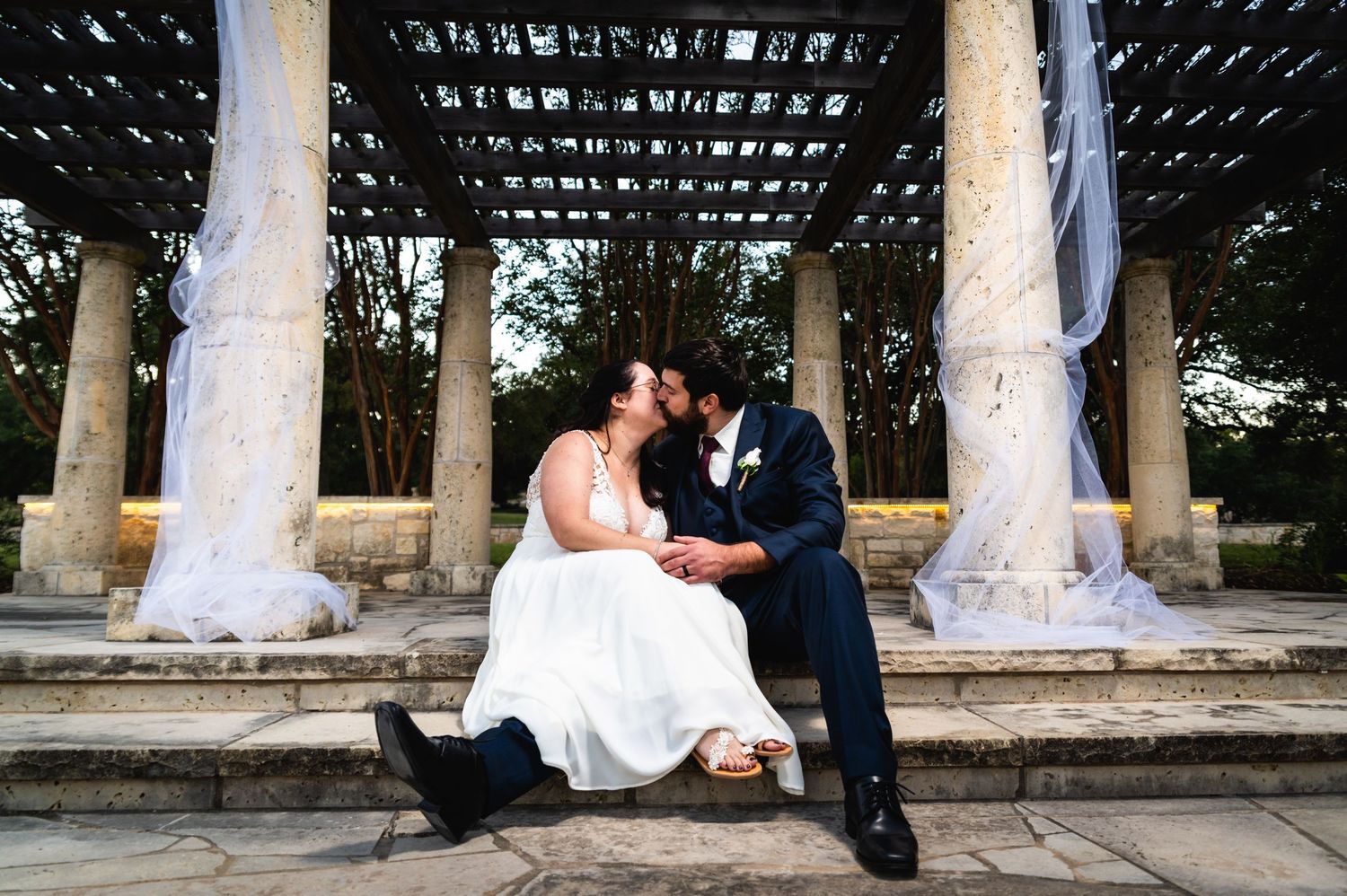 Romantic wedding couple sharing a moment on stone steps under a latticed pergola.