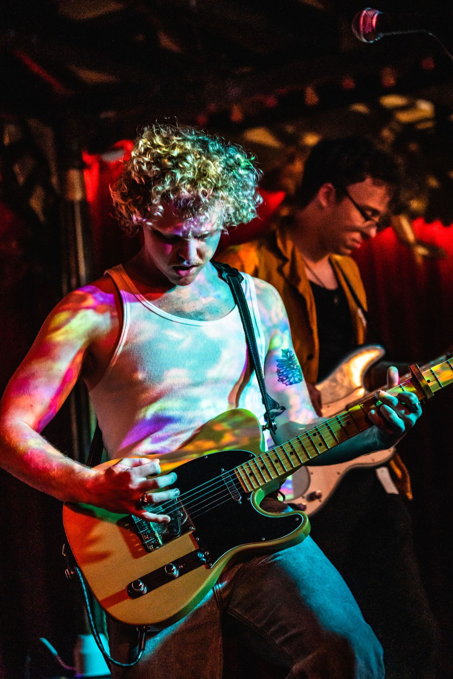 Musician with curly hair playing guitar under colorful stage lights.