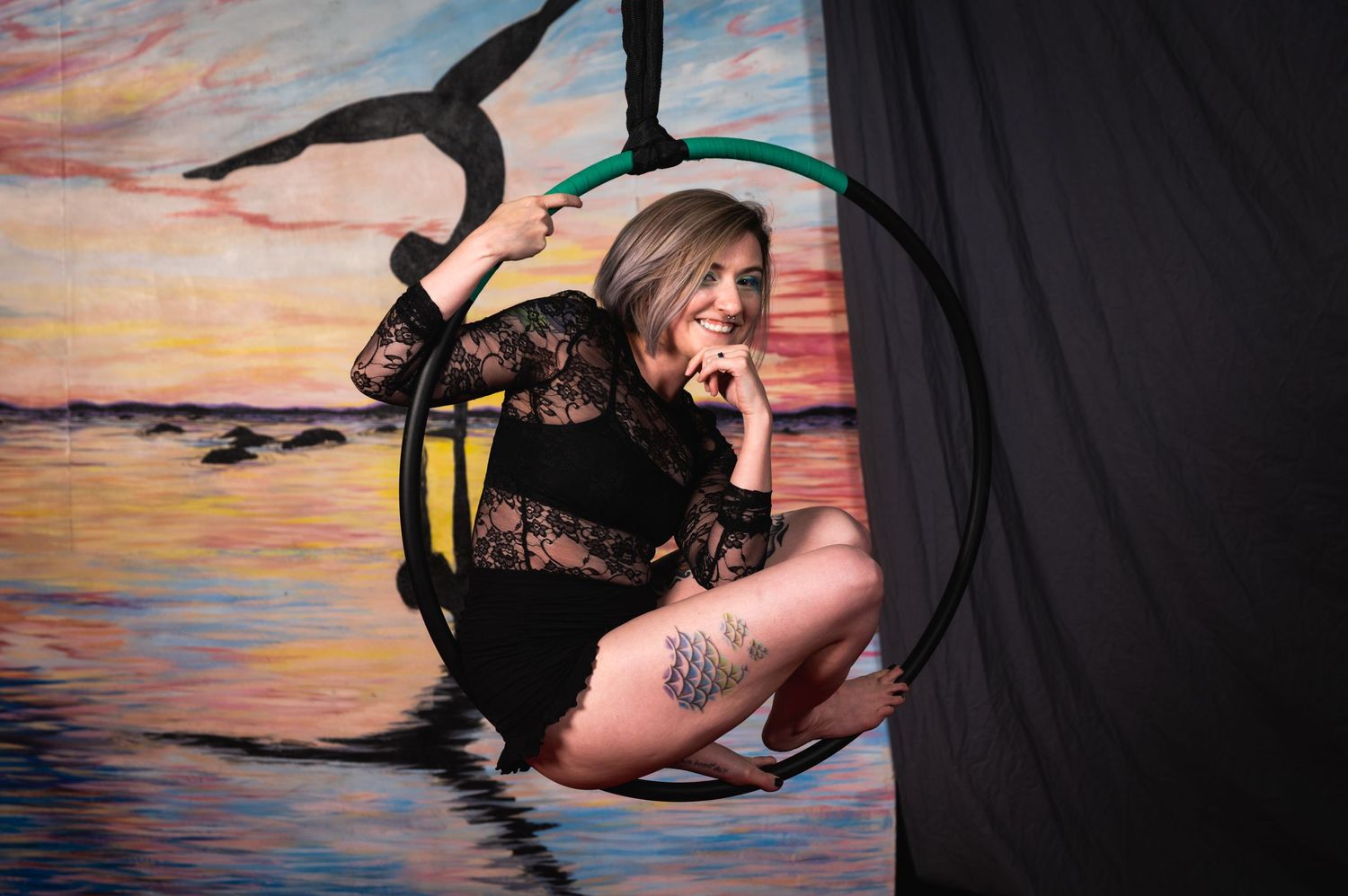 Aerial artist in black lace performs on green hoop against painted sunset background.
