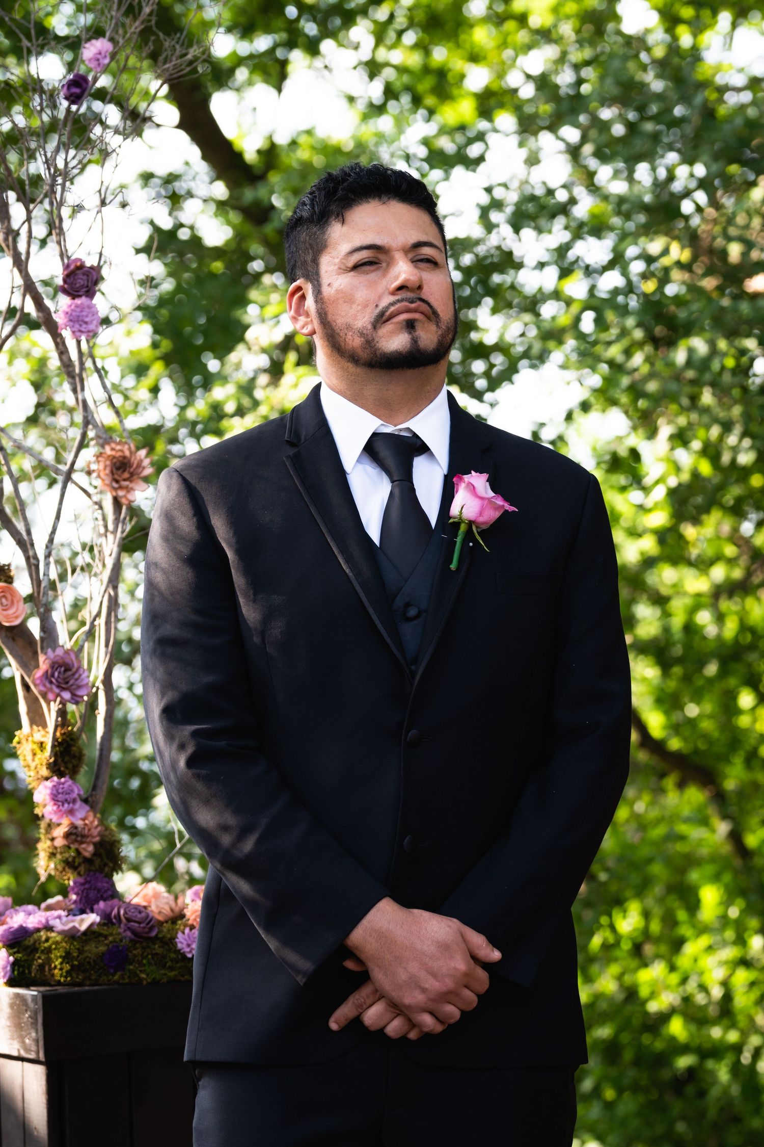 A groom in a black tuxedo with pink boutonniere stands at the wedding altar.