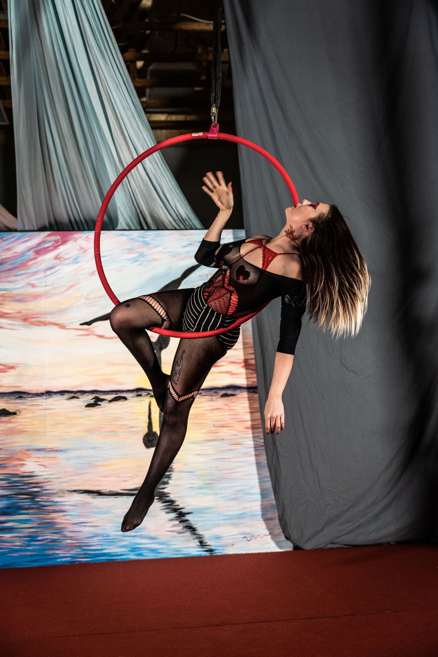 An aerial performer in a black and red costume gracefully poses with a red hoop against a sunset backdrop.