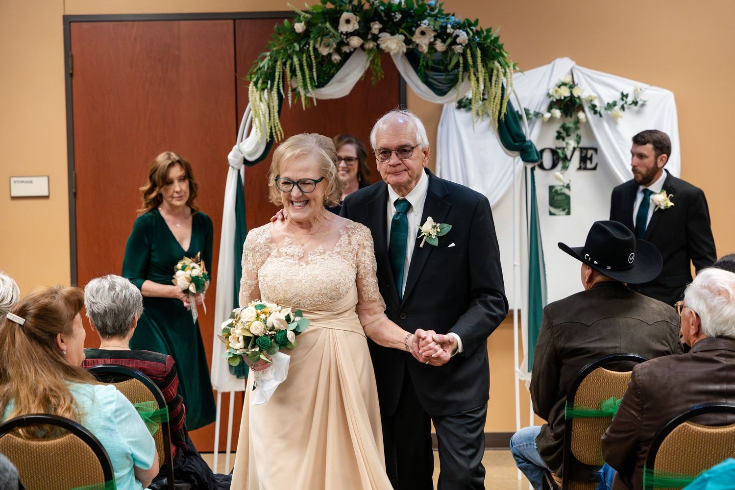 A couple walks down the aisle under a floral arch during a wedding ceremony while guests look on.