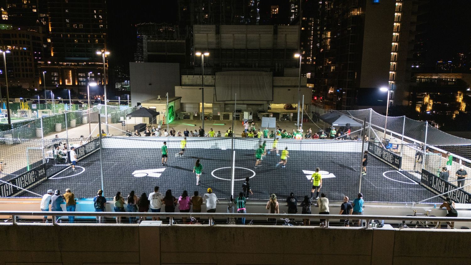 Aerial view of an illuminated urban soccer court at night with spectators watching from the sidelines.