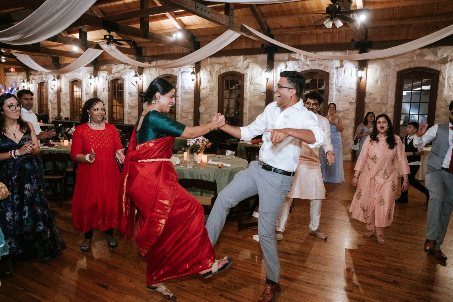 Party guests dance energetically on a wooden dance floor at an indoor reception venue.