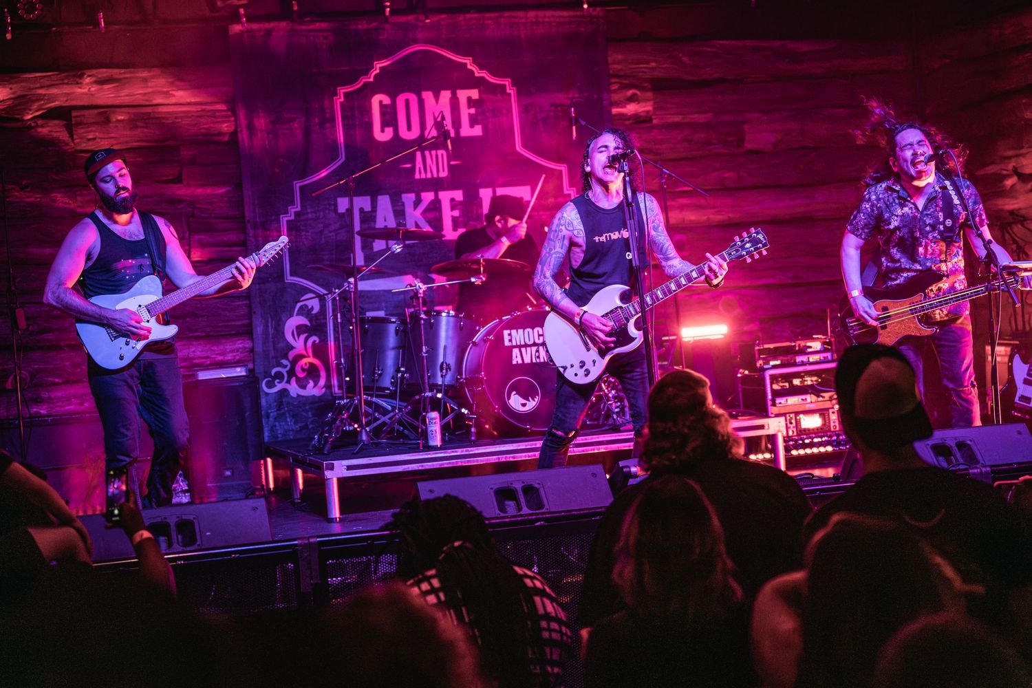 Rock band performs on stage with multiple guitarists under red lighting at a live music venue.