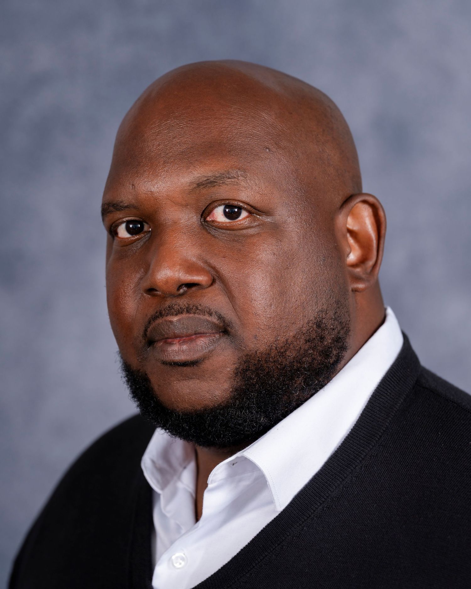 Professional headshot portrait showing a confident person wearing a white dress shirt and black blazer against a gray background.