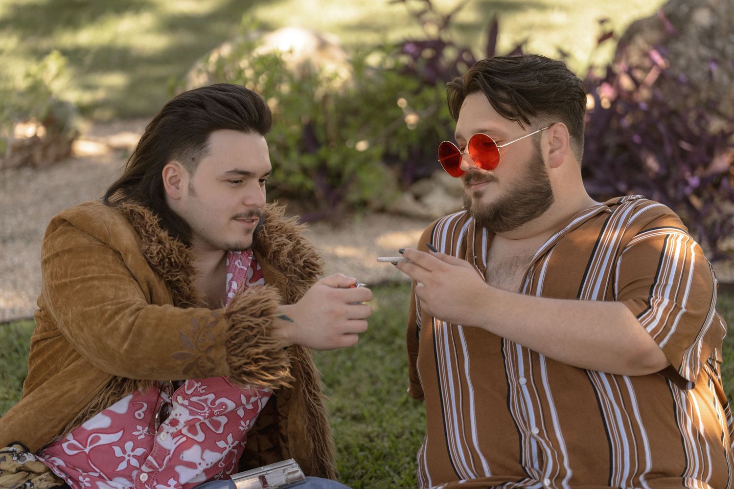 Two people in vintage-inspired outfits share a lighter in an outdoor garden setting.