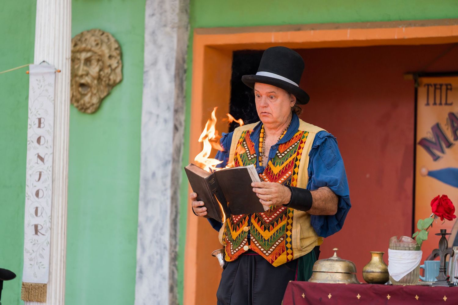 Street performer in traditional vest and top hat performs with flaming book at outdoor festival.