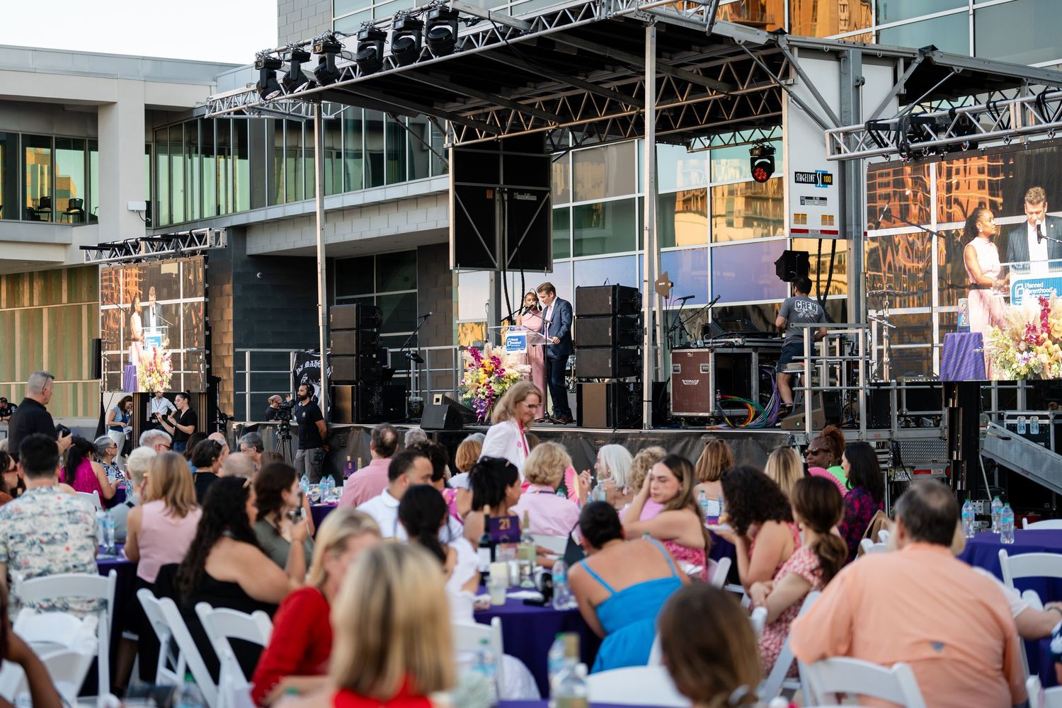 Outdoor event setup with stage equipment and seated audience at tables during sunset.