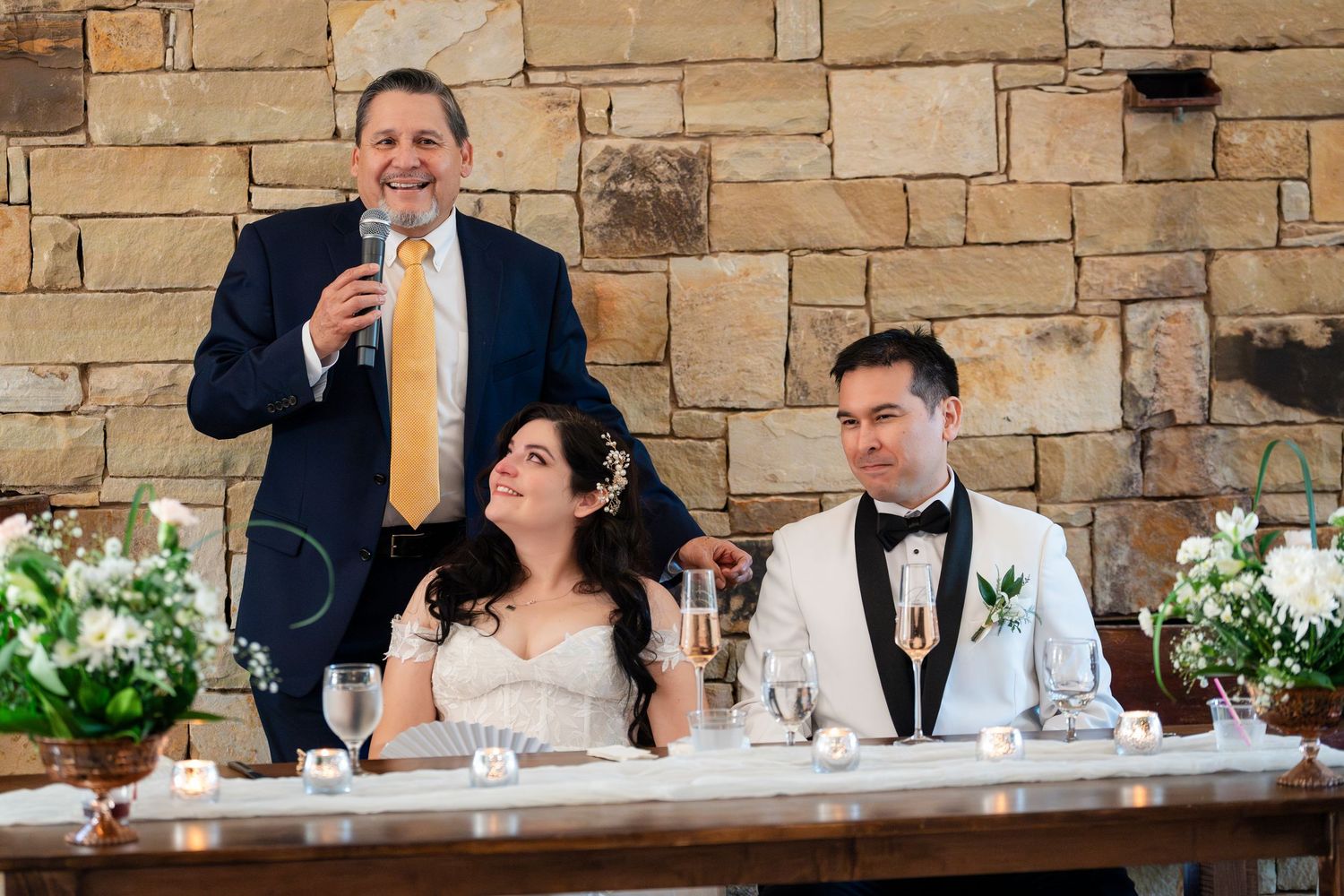 Wedding scene at a stone wall venue with guests in formal attire seated at an elegant decorated table.