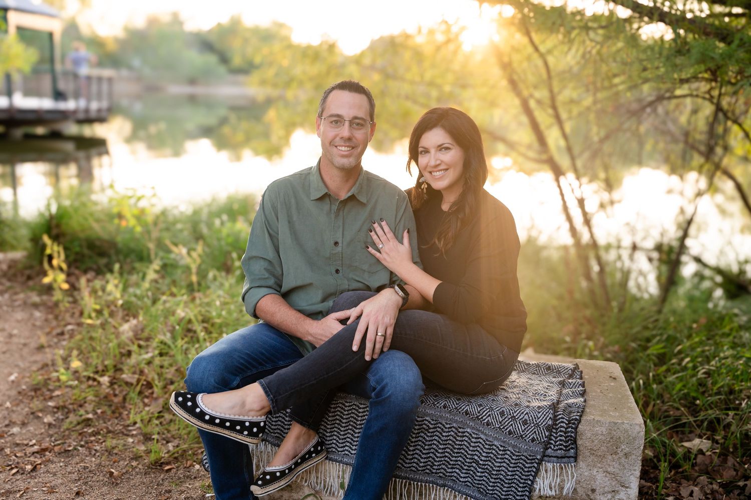 A couple sits together on a woven bench by a lake during sunset.