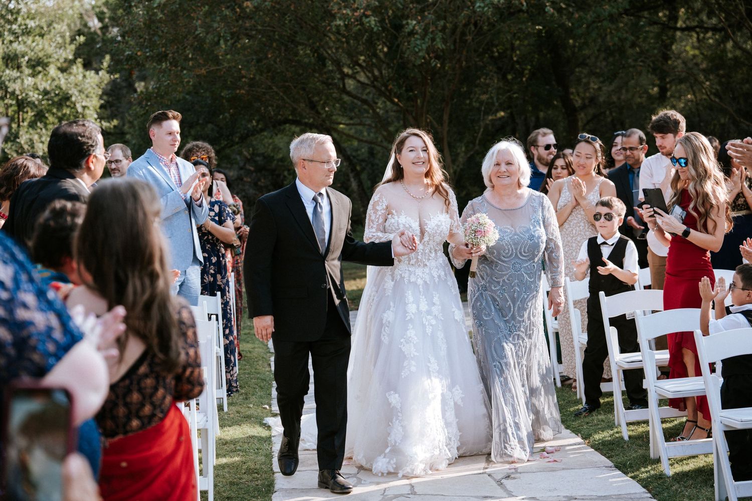 Wedding ceremony guests watch as the bride in lace gown walks down an outdoor aisle with family members.
