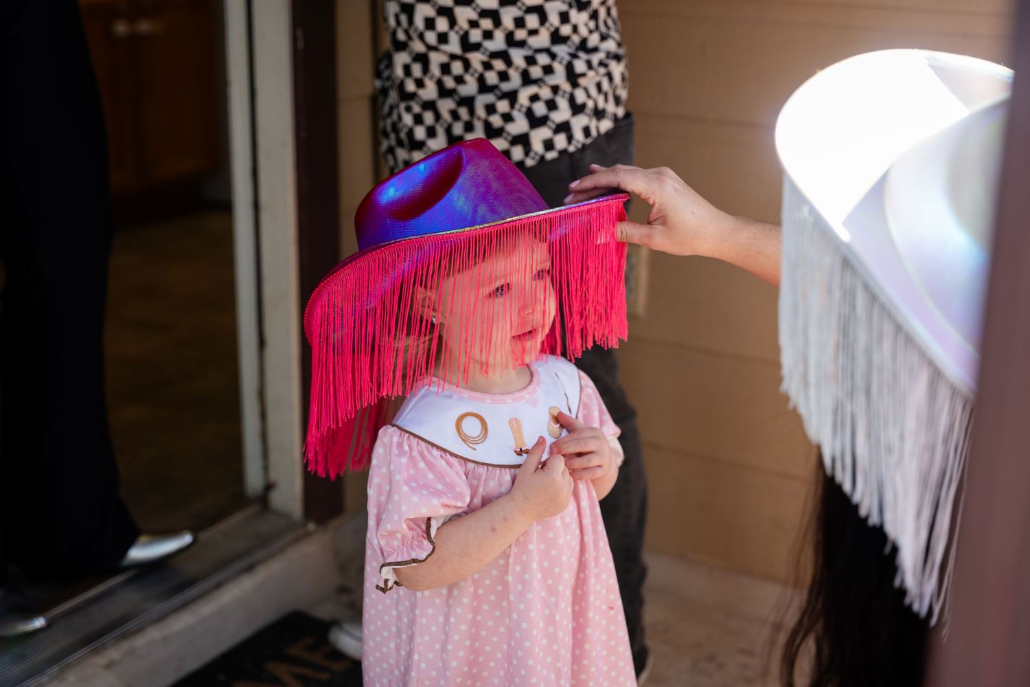 A little one wearing a pink dress plays dress up with a bright blue hat with pink fringe tassels.