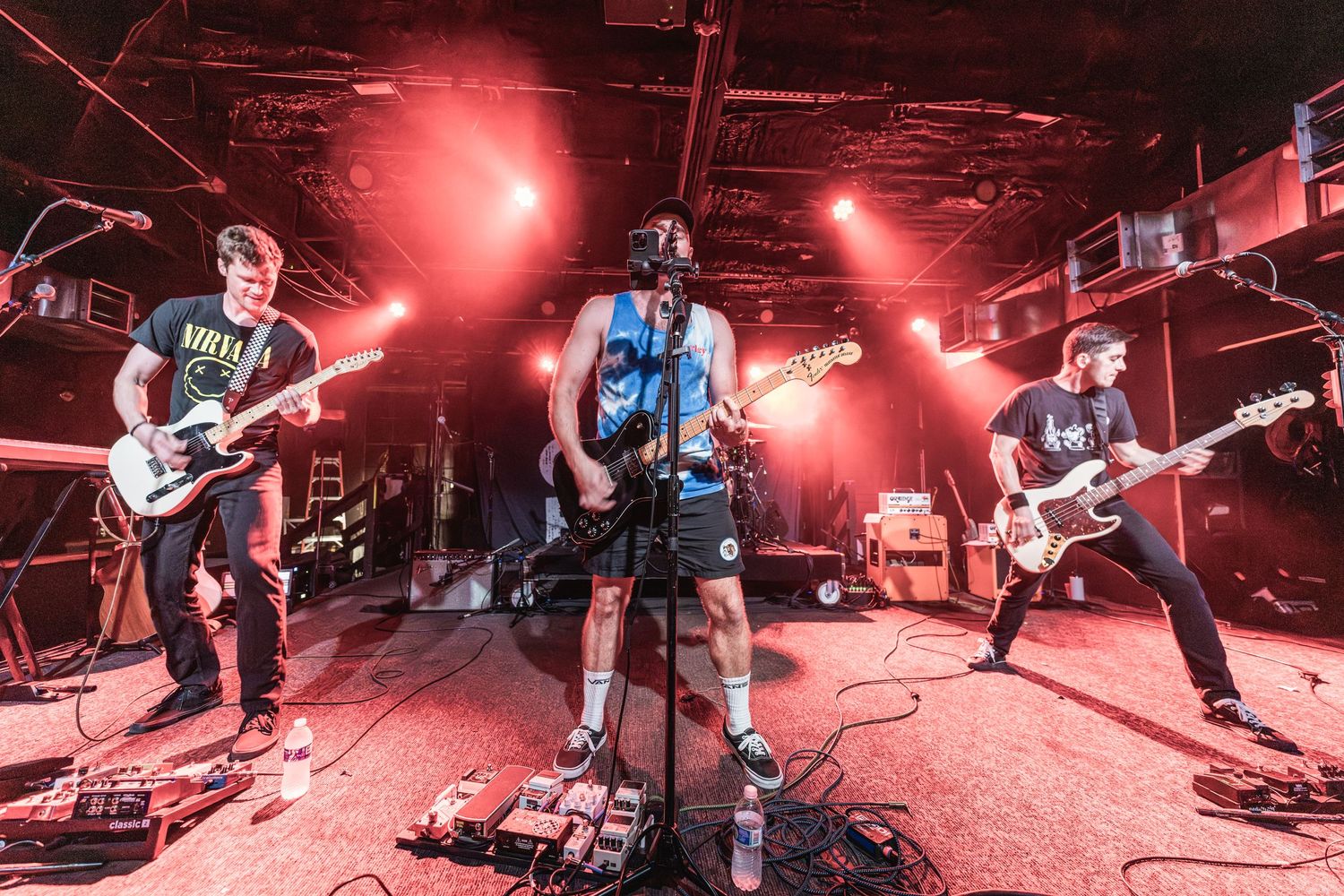 Rock band performs on stage under dramatic red lighting with guitars and microphones.