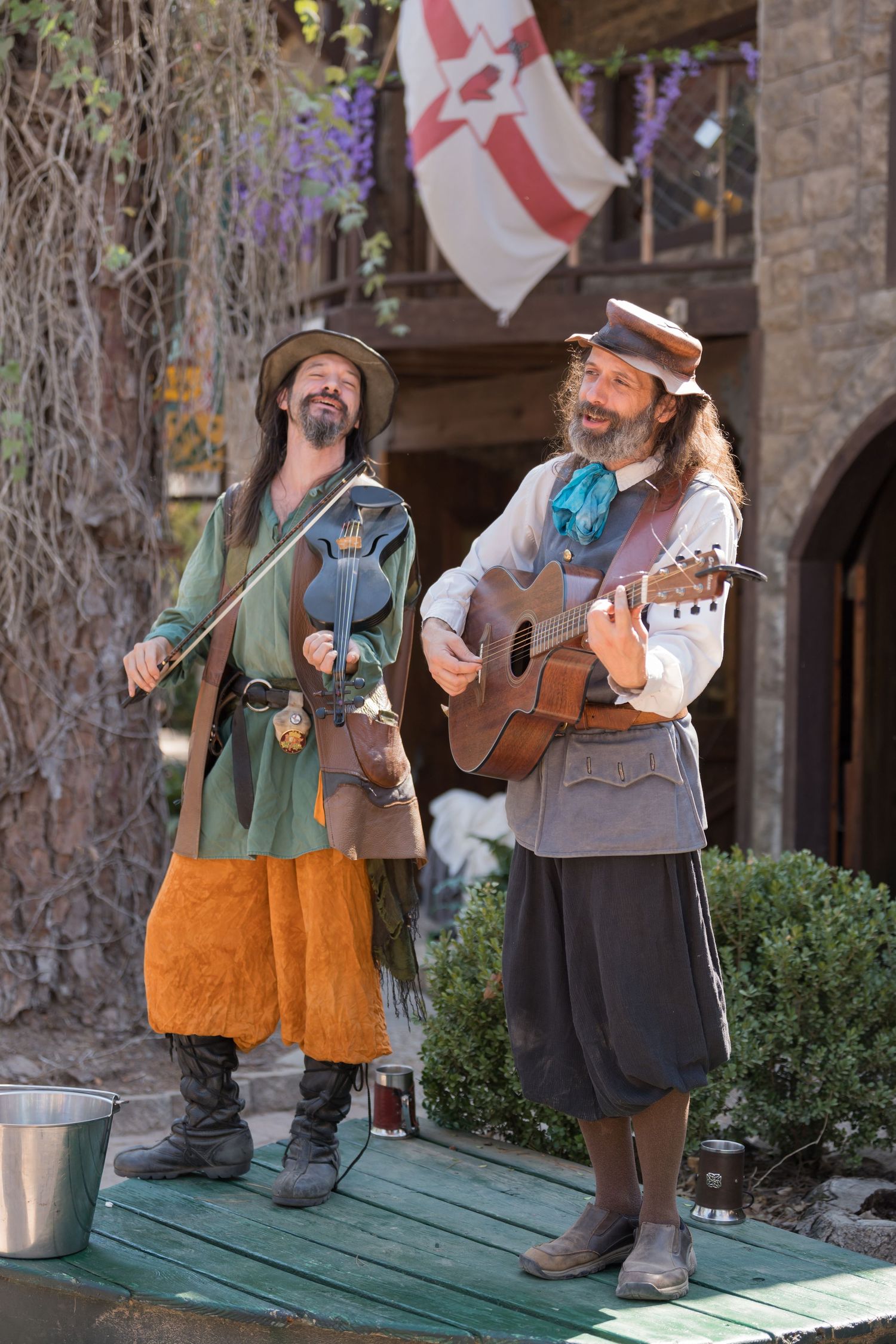 Two medieval-style musicians perform on a wooden stage with a flag backdrop at a renaissance faire.