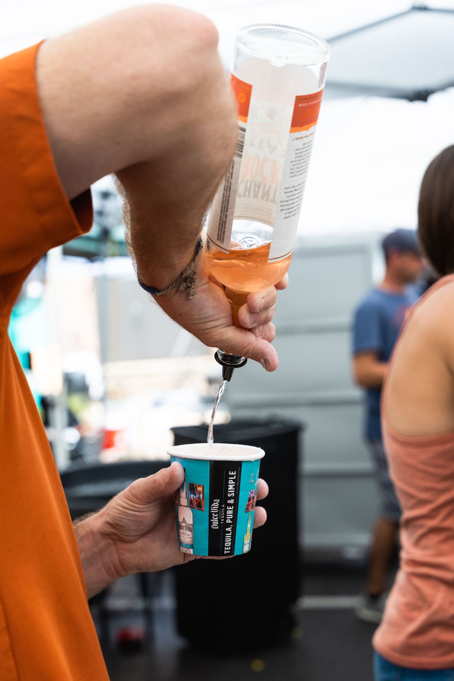 Bartender pours a measured drink from a bottle into a small plastic cup at an outdoor event.